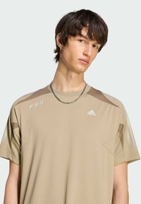 Béžové sportovní tričko s krátkými rukávy, s texturovaným vzorem na ramenou a logy na hrudi a rameni v kontrastních barvách.