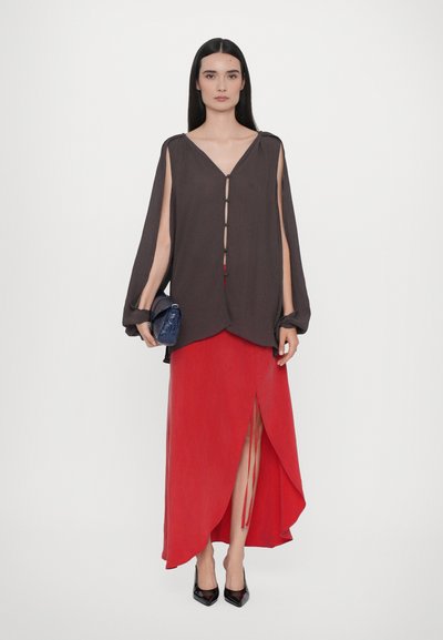 Braunes Langarmtop mit V-Ausschnitt und Knopfverschluss, kombiniert mit einem roten, knöchellangen Rock mit seitlichem Schlitz und Bindedetail. Schwarze High Heels.