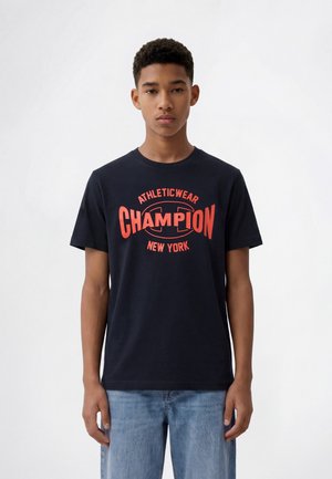 Jeune homme portant un t-shirt noir avec un texte rouge "Athleticwear Champion New York", associé à un jean bleu clair, debout devant un fond blanc.