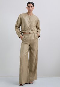 Chaqueta corta beige con dobladillo elástico y pantalones de pierna ancha a juego, ambos con patrones fluidos y acentos metálicos. Textura de tela suave.