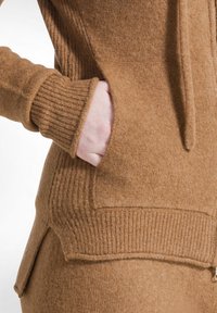Ruskea neulehuppari, jossa on ribbauskohdat hihansuissa ja helmassa, sivutaskut ja teksturoitu kangaspinta. Muotoilu korostaa lämpöä ja mukavuutta.