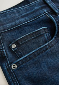 Dark blue denim fabric featuring a front pocket with visible stitching and a metal button accent. Smooth texture and structured shape.