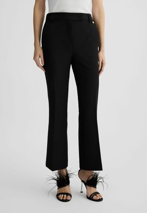 Black tailored trousers with a straight cut and plain texture. Worn with black strappy heels featuring feather accents at the ankles.