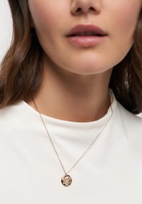 Halskette aus Roségold mit einem runden Anhänger, der florale Gravuren aufweist. Der Anhänger hängt an einer zarten Kette, die zu einem weißen Oberteil passt.