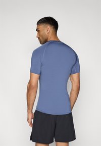 Αθλητική μπλούζα με κοντά μανίκια σε μπλε χρώμα, κατασκευασμένη από ελαστικό ύφασμα. Διαθέτει εφαρμοστή γραμμή, επίπεδες ραφές και σκούρο λογότυπο της μάρκας στο λαιμό.