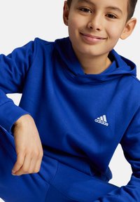 Felpa blu in cotone con cappuccio regolabile, caratterizzata da un logo Adidas bianco sul petto e polsini a coste. Texture liscia e vestibilità comoda.