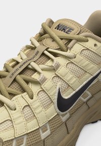 Μπεζ αθλητικό παπούτσι Nike με πάνελ από διχτυωτό ύφασμα και δέρμα, μαύρο κεντημένο σήμα swoosh, χοντρή σόλα και κορδόνια που περνούν μέσα από οριζόντιους βρόχους.