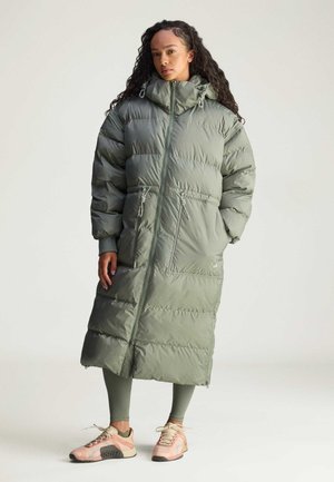 Een vrouw staat in een lange, groene pufferjas met een capuchon, gecombineerd met strakke leggings en stijlvolle sneakers, tegen een minimalistische achtergrond.