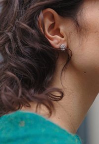 Brincos redondos de prata com um padrão texturizado, apresentados em um design de aro. Cabelo encaracolado emoldura a orelha, com textura de pele visível e uma peça de vestuário azul-esverdeada.