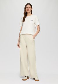 T-shirt en coton blanc avec un graphique en forme de cœur sur la poitrine gauche. Pantalons larges crème avec poches latérales. Le mannequin porte des baskets au design crème et noir.