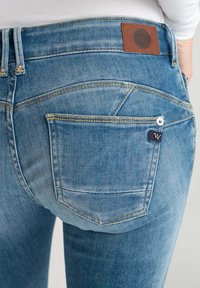 Jean en denim bleu avec une finition délavée, featuring une pièce en cuir marron, deux poches arrière et des détails de couture jaune.