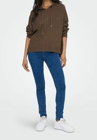 Brun, ribbad knit hoodie med dragsnoddar, oversized passform. I kombination med åtsittande blå jeans och vita sneakers, vilket skapar en avslappnad, lager-på-lager-look.