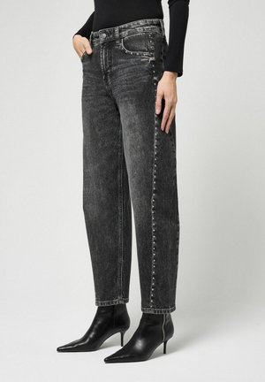 Vrouw draagt hoog getailleerde vervaagde zwarte jeans met kleine studs langs de zijnaden en zwarte puntige enkellaarsjes met hak, staand tegen een effen achtergrond.