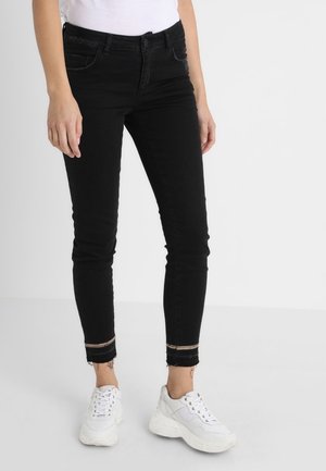 Femme portant un jean skinny noir avec des ourlets effilochés et un détail rayé, associé à des baskets blanches épaisses et un haut blanc.