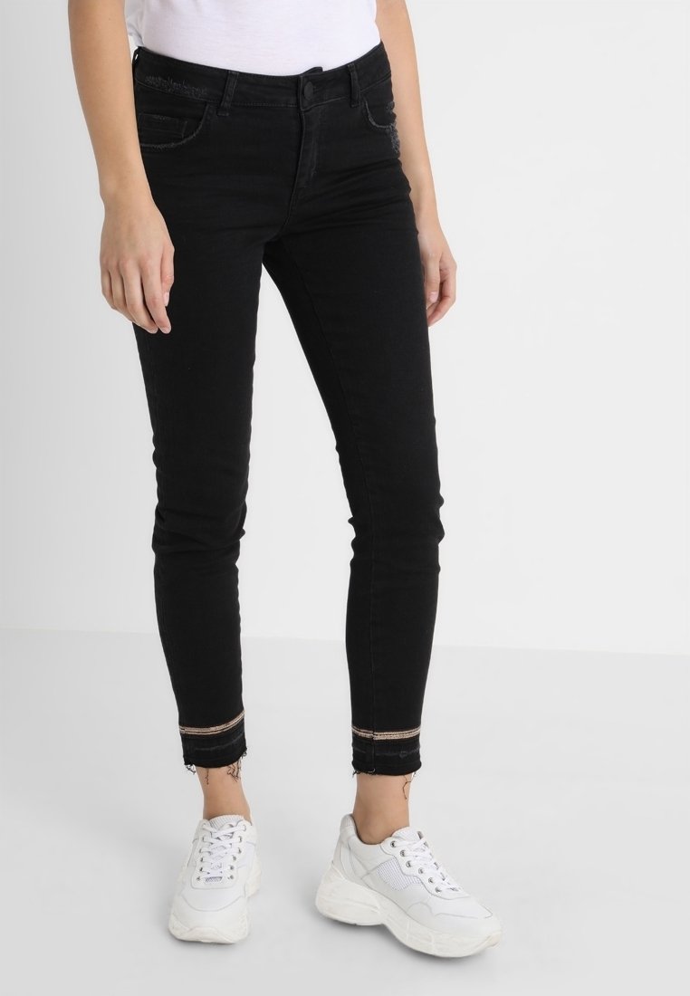 Femme portant un jean skinny noir avec des ourlets effilochés et un détail rayé, associé à des baskets blanches épaisses et un haut blanc.