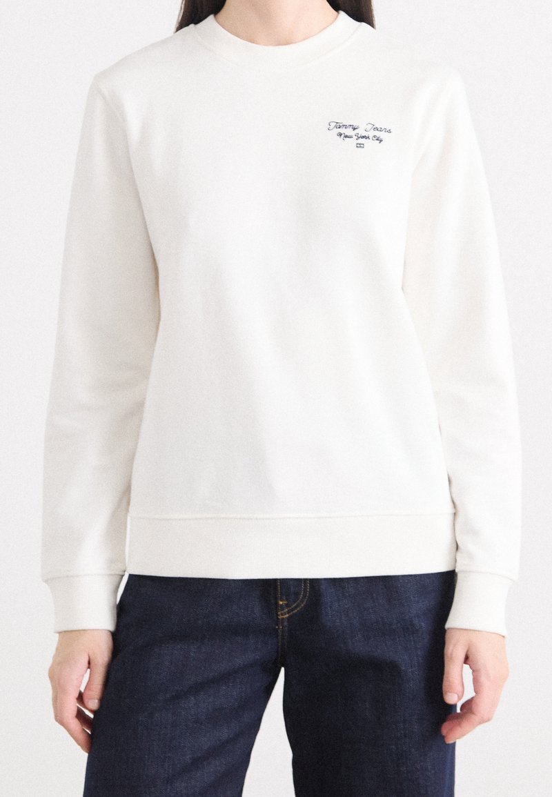 Sweat-shirt blanc à manches longues avec petit texte noir "Tommy Jeans New York City" sur la poitrine gauche, porté avec un jean bleu foncé.