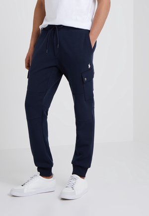 Mann trägt dunkelblaue Jogginghose mit seitlichen Cargotaschen und weißen Sneakers, steht mit Händen in den Taschen vor einem schlichten Hintergrund.