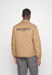 Chaqueta acolchada en tonos marrones con cuello alzado, que presenta letras en negro en la parte posterior y una superficie texturizada. Combinada con pantalones grises.