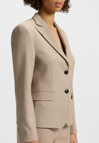 Blazer aderente beige realizzato in tessuto testurizzato, con rever a punta, due bottoni neri e tasche laterali, progettato per una silhouette strutturata.