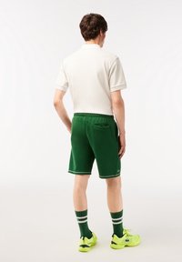 Man som bär en vit polo-shirt, gröna shorts, gröna strumpor med vita ränder och ljusgula sneakers, stående vänd bort från kameran mot en vit bakgrund.