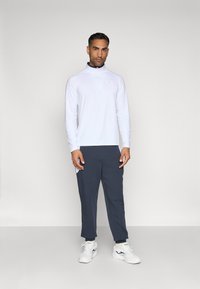 Balts garo piedurkņu džemperis ar rāvējslēdzēja apkakli, kopā ar tumši ziliem biksēm un baltiem sporta apaviem. Minimālistisks dizains, gluda auduma tekstūra.