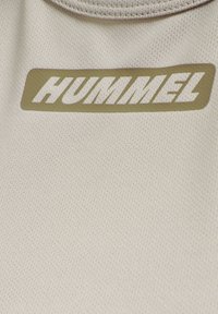 Beige textuurstof met een taupekleurig rechthoekig logo met het woord "HUMMEL" in hoofdletters dicht bij een ronde halslijn.