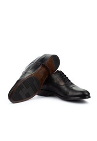Zapatos de vestir para hombre de cuero negro con cordones, uno mostrando la suela pulida con patrones de agarre, exhibido sobre un fondo blanco.