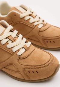 Zapatillas de ante marrón claro con cordones crema, que presentan una punta redondeada, detalle de costuras y pequeños orificios de ventilación en los laterales.