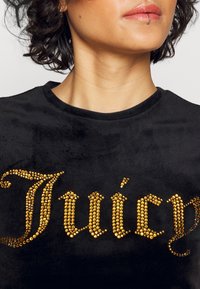 Svart sammet t-shirt med en stor "Juicy" design utsmyckad med gyllene rhinestones, som visar en texturerad yta och rund halsringning.