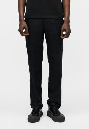 Homme portant un pantalon noir droit, des baskets noires à lacets et une chemise noire, debout devant un fond uni.