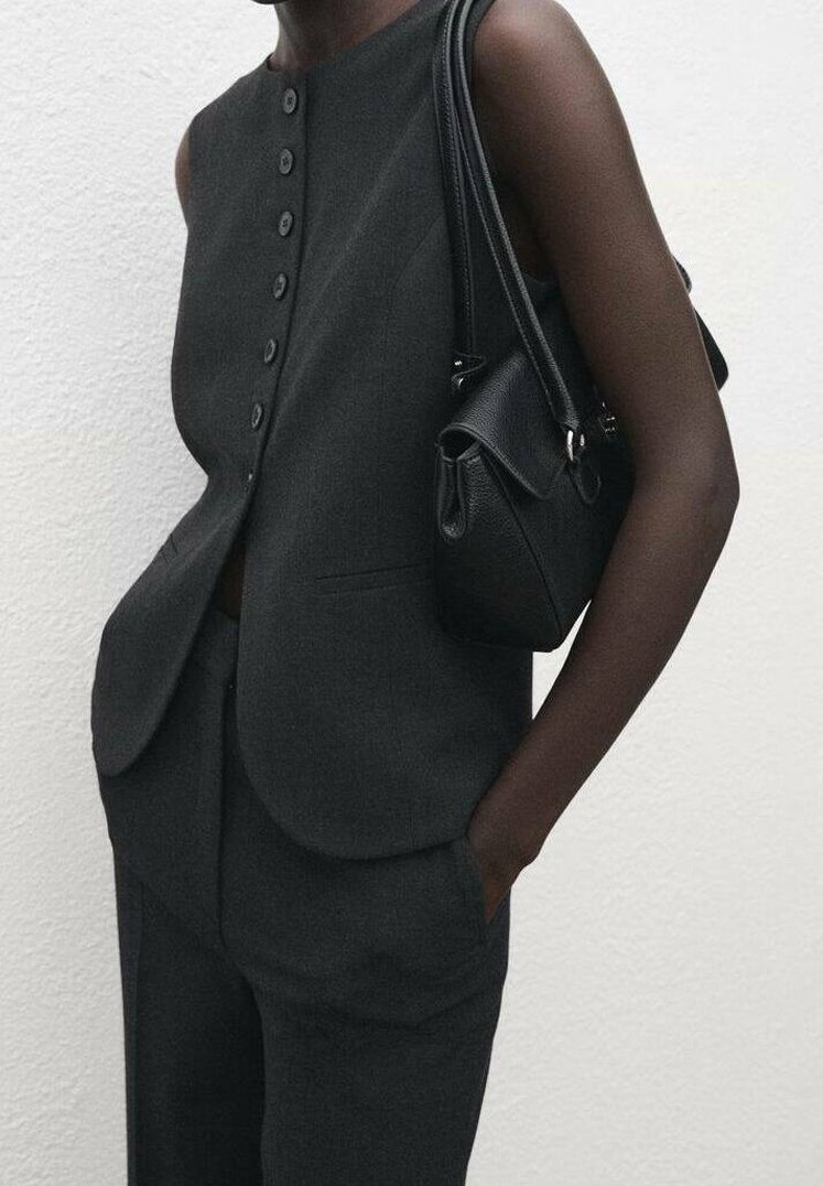 Personne portant un gilet sans manches gris foncé boutonné et un pantalon assorti, une main dans la poche et un sac à main noir à l'épaule.