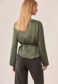 Blusa verde oliva con un acabado texturizado, mangas largas y cintura fruncida, combinada con unos vaqueros negros de cintura alta.