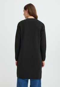 Vêtement tricot noir à manches longues avec une coupe droite, doté d'un col rond et de manches tombantes, atteignant le milieu de la cuisse.
