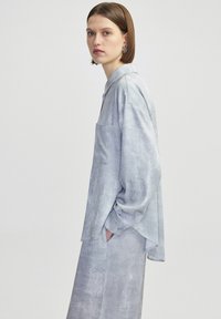 Camisa de botones de color gris claro con mangas largas, un bolsillo en el pecho y un ajuste ligeramente oversize; confeccionada en una tela suave y texturizada.