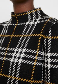 Gros plan sur un pull noir à motif à carreaux blanc et jaune porté par une personne à la peau foncée.