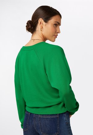 Tummatukkainen nainen, jolla on matala nuttura, kultaiset rengaskorvakorut, vihreä neulepusero ja siniset farkut, katsoo olkansa yli valkoista taustaa vasten.