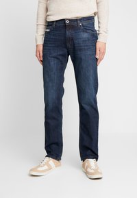 Jeans de mezclilla azul oscuro con corte recto, que cuentan con cinco bolsillos, cierre de cremallera y un sutil detalle de desgastado, combinados con zapatillas beige y blancas.