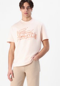 Hellrosa Baumwoll-T-Shirt mit einem grafischen Druck eines Krokodils und dem Markennamen "Lacoste" in Orange. Kurze Ärmel und runder Halsausschnitt.