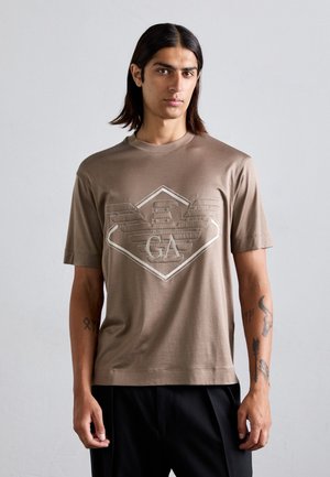 Jonge man met lang, donker haar draagt een bruine T-shirt met een reliëfafbeelding van een adelaar en de letters "GA", gecombineerd met zwarte broek, staand voor een effen achtergrond.