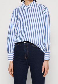 Camisa abotonada de rayas azules y blancas en tejido ligero, con un bolsillo en el pecho y mangas largas con puños de rayas en contraste.