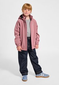 Helle rosa Softshell-Jacke mit Kapuze, graues T-Shirt darunter, lockere dunkle Jeans und blaue Sneakers mit weißen Akzenten. Sichtbare Sommersprossen.