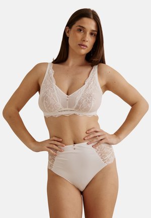 Femme posant les mains sur les hanches, portant un soutien-gorge en dentelle rose clair et des culottes taille haute, sur fond uni.