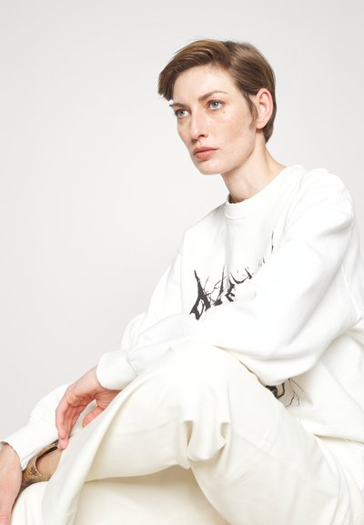 Λευκή βαμβακερή μπλούζα με μαύρο γραφικό σχέδιο, συνδυασμένη με φαρδιά λευκά παντελόνια. Η λεία υφή και η oversized εφαρμογή είναι προφανείς.