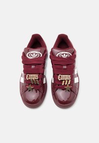 Ζευγάρι γυαλιστερών μπορντό αθλητικών παπουτσιών Adidas Campus με λευκές ρίγες, μπορντό κορδόνια και χρυσά διακοσμητικά "Campus" και αστέρια στα κορδόνια.