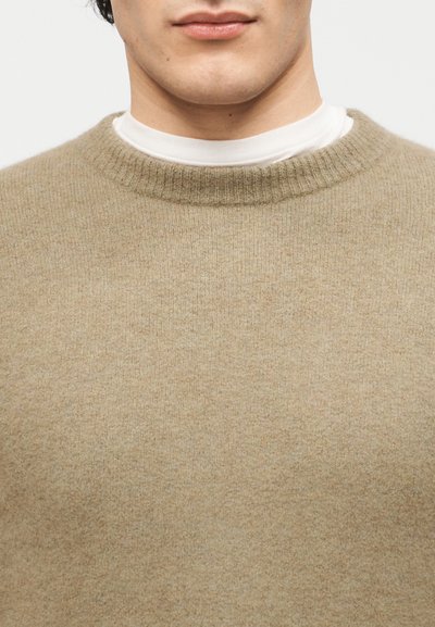 Άνδρας που φοράει ανοιχτά καφέ πουλόβερ με στρογγυλή λαιμόκοψη πάνω από λευκό πουκάμισο με ψηλό γιακά, κοντινή όψη από τον λαιμό μέχρι το πάνω μέρος του στήθους.