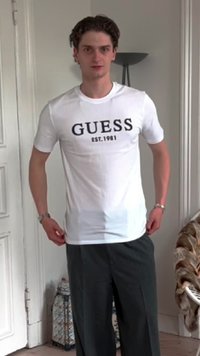 Bela kratka majica s kratkimi rokavi z črnim napisom "GUESS EST.1981" na sprednji strani. V paru s sivo nagubano hlačami, stoji ob nevtralni ozadju.