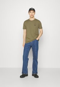 Cămașă verde măsliniu cu un mic logo, blugi negri din denim și pantofi negri, purtate cu o șapcă neagră. Design simplu și croială relaxată.