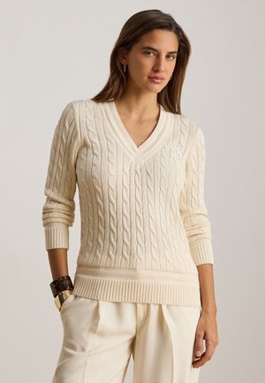 Kvinde iført cremefarvet kabelstrikket V-hals sweater med lange ærmer og cremefarvede bukser med høj talje, stående med den ene hånd i lommen.