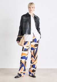 Chaqueta corta de cuero negro con grandes bolsillos, camisa blanca de botones, pantalones de pierna ancha de colores con un patrón en naranja, azul y marrón, zapatos negros, bolsa beige.