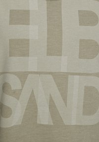 Beige T-särk, millel on suured, kulunud tähekirjad heledas valges kuues tekstuurses materjalis. Kujunduses on hõredad ja tumedad värvimuutused.